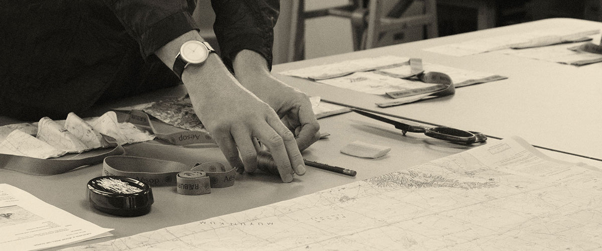 Hands at a cutting table measuring out reclaimed navigation map fabric and Aesop × RÆBURN tape for the RÆMADE Adventurer Roll Up.