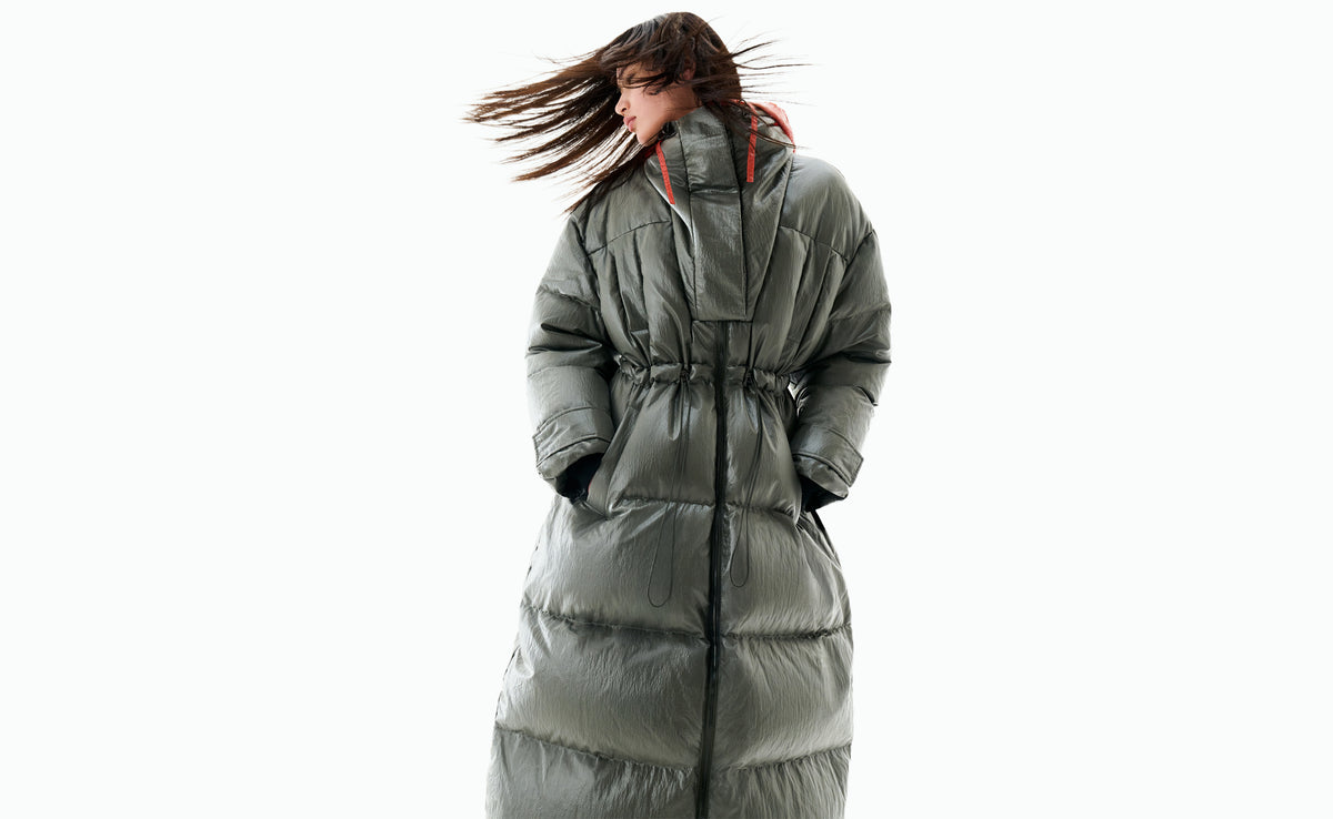 A model with wind-blown hair in a long khaki down coat with orange drawcords at the collar.