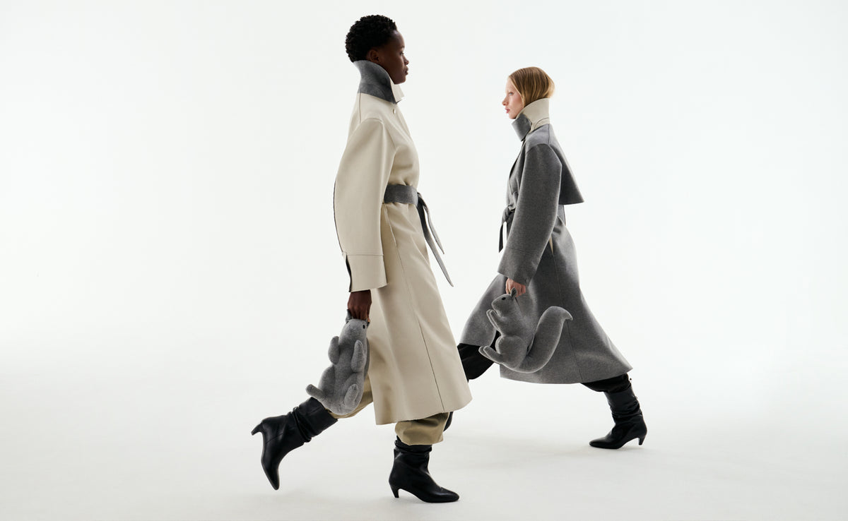 Two models walking in profile in two-tone reversible wool coats, one in stone and one in grey, each carrying the collection’s grey squirrel mascot.
