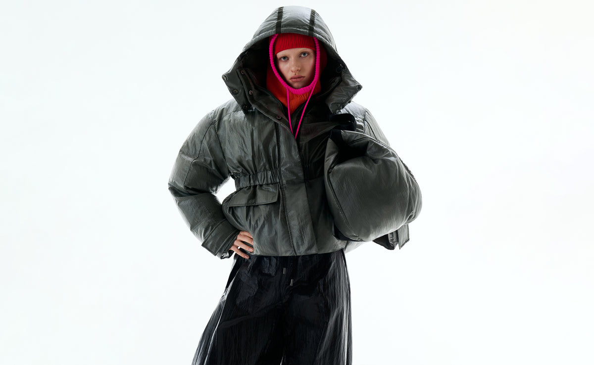 A model in a hooded khaki down jacket and an orange balaclava with pink trim, holding a khaki maxi bag under one arm.