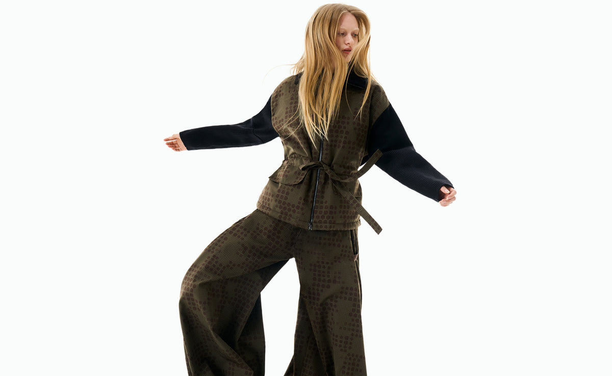 A model mid-movement in a belted khaki jacquard jacket with black knit sleeves and matching wide-leg trousers.
