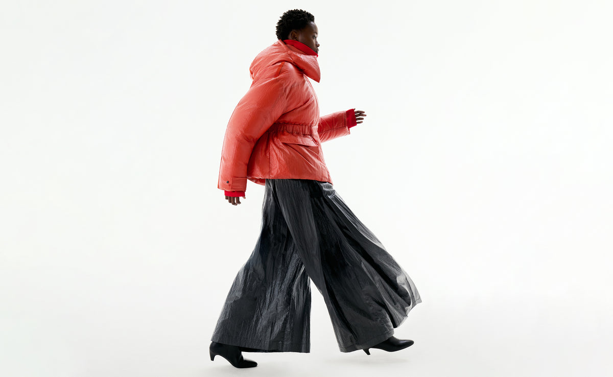 A model walking in profile in an orange down jacket and wide black trousers.