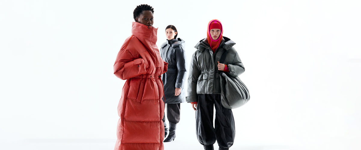 Three models in MAX&Co. × RÆBURN outerwear: a long orange down coat, a dark grey padded coat, and a khaki down jacket with an orange balaclava and maxi bag.