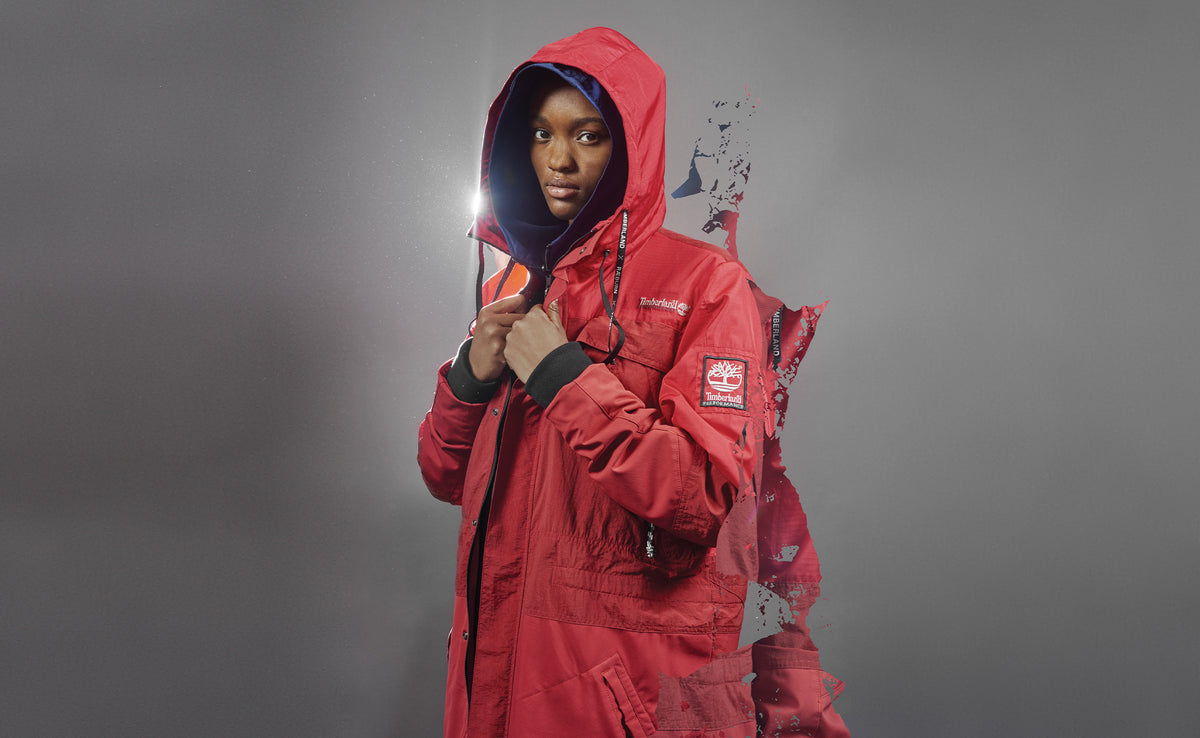 Model in a red hooded Timberland × RÆBURN parka, lit from behind against a grey backdrop.